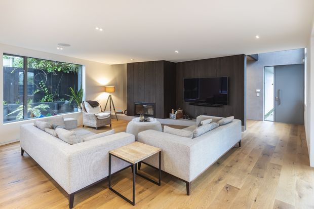 A bright, spacious contemporary living room with two large light upholstered sofas arranged around a low coffee table and side table. The room features wide-plank timber flooring, a dark timber feature wall with built-in fireplace and wall-mounted television, recessed ceiling lights, and large floor-to-ceiling glazing overlooking a planted courtyard. A grey tiled or concrete-toned entry zone is visible at the right.