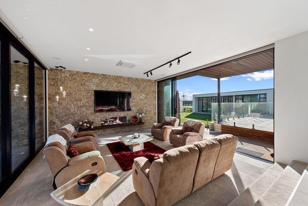 A bright, spacious contemporary living room with multiple tan upholstered sofas and armchairs arranged around a low coffee table and deep red rug. A stone-clad feature wall incorporates a wall-mounted television and linear fireplace. Full-height glazed sliding doors open to a covered outdoor terrace with timber-lined soffit and views toward a modern secondary structure. A stair edge is visible in the foreground.