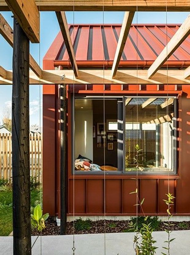 Exterior view of a compact red metal-clad dwelling or extension, seen through a timber pergola or exposed post-and-beam structure. The facade has a steep standing-seam metal roof, large glazed openings with dark frames, and a visible bedroom or sleeping area through the open window. A paved path and planted garden border the building.