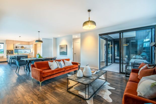 Open-plan contemporary living space with a warm-toned timber floor, white walls, and large dark-framed sliding glass doors. A vivid rust-orange sofa grouping surrounds a black-framed glass coffee table, while the adjoining dining and kitchen area is visible in the background. Pendant lights, built-in cabinetry, artwork, and a textured hide-style rug complete the polished interior.