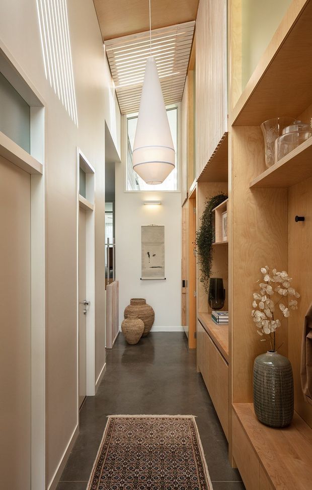 A narrow, refined residential hallway with a dark smooth floor, pale walls, and extensive light timber built-in shelving and cabinetry on the right. The corridor features a tall glazed opening, a slatted timber ceiling detail filtering daylight, a large sculptural pendant light, recessed wall niches, pottery, decorative objects, and a patterned runner rug. The space has a warm, minimalist contemporary character with strong vertical proportions and abundant natural light.