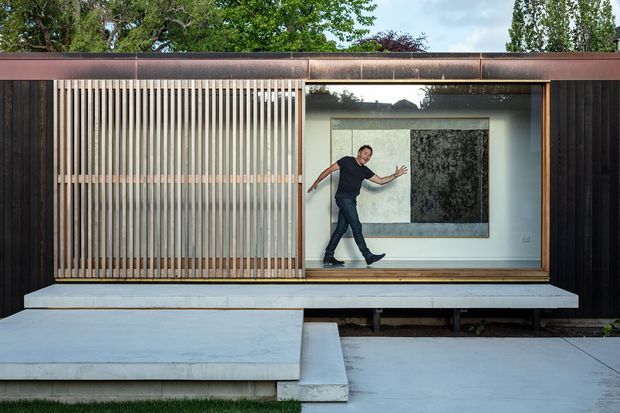 A contemporary single-storey building facade with dark vertical cladding, a broad rust-toned metal roof edge, and a large glazed opening. Vertical timber privacy screens flank the glazing, while a pale concrete platform and stepped entry extend across the foreground. Through the window, a person and large monochromatic artwork are visible inside. Mature trees appear behind the building.