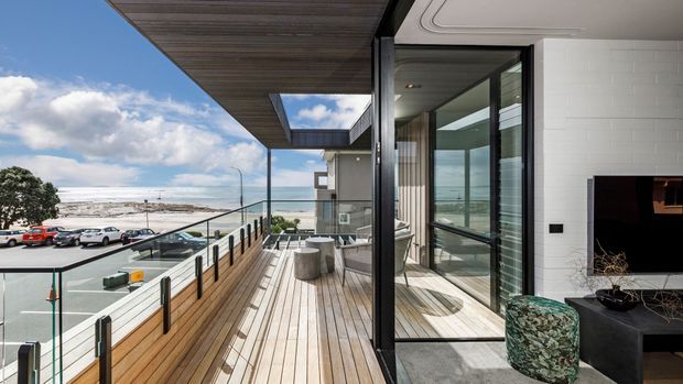 A contemporary coastal apartment terrace or balcony overlooking the ocean. The space features timber decking and soffit lining, clear glass balustrades with dark metal framing, extensive floor-to-ceiling glazing, and a white masonry wall with a wall-mounted television. Outdoor seating and a small side table occupy the sheltered terrace, with a coastal street and shoreline visible beyond.