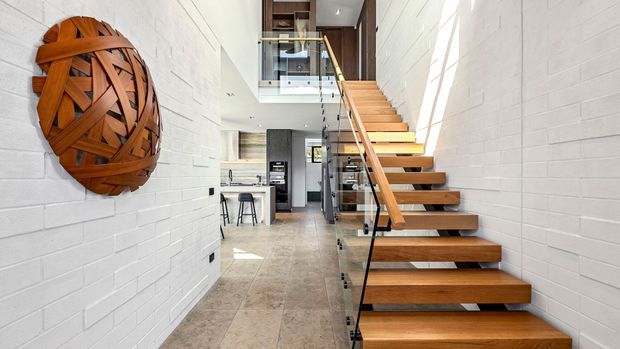 Bright contemporary interior focused on an open timber staircase with glass balustrades, set between white-painted brick walls. A compact kitchen and mezzanine-level landing are visible beyond, while large-format stone-look floor tiles and exposed stair treads create a clean, airy vertical circulation space.