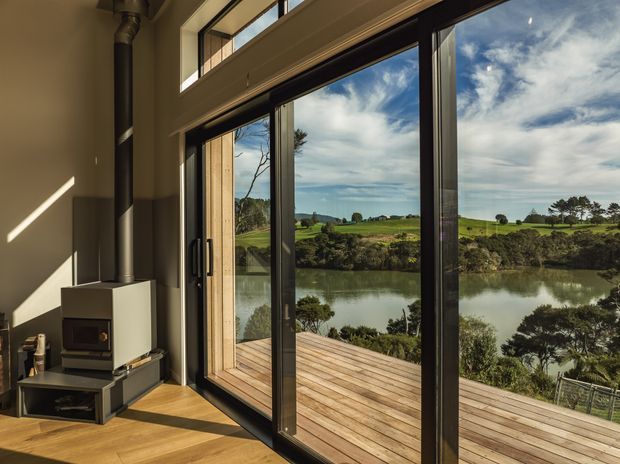 A bright contemporary interior opening onto a timber deck through large black-framed sliding glass doors. The room contains a compact freestanding wood-burning stove with a dark hearth, warm timber flooring, and high glazing above the doors. Beyond the deck is a calm waterway and green rural landscape under a partly cloudy sky.