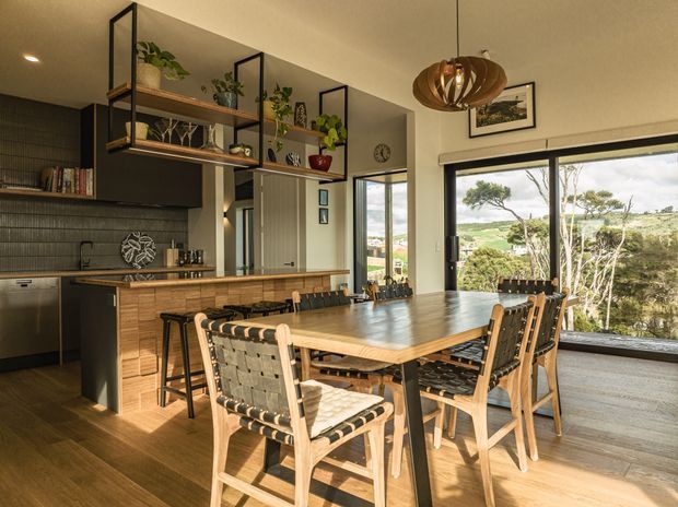 A warm, open-plan kitchen and dining area with dark cabinetry, timber flooring, a timber kitchen island, and a large rectangular dining table surrounded by woven timber chairs. Open black-framed shelving displays plants and decor, while expansive glazing brings in abundant daylight and views of greenery outside. A sculptural timber pendant light hangs above the dining table.