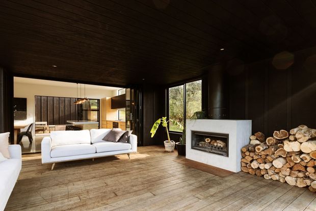 A warm, dark-toned contemporary living room with a white sofa, timber flooring and a full-height glazed opening to an adjoining dining and kitchen area. The room features a pale stone or plaster fireplace surround with an inset firebox, stacked firewood, dark timber wall and ceiling lining, and large windows overlooking greenery.
