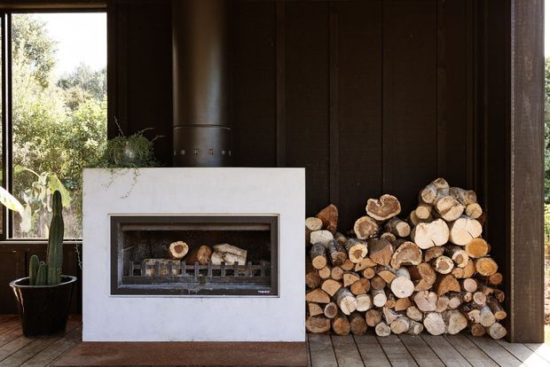 A dark, timber-lined interior centered on a white rectangular fireplace with a built-in firebox. A tall black flue rises above the mantel, while a stacked woodpile occupies the adjacent wall. Large windows reveal greenery outside, and potted cacti sit near the fireplace. The space has a warm, rustic cabin-like character with strong natural materials and high contrast.