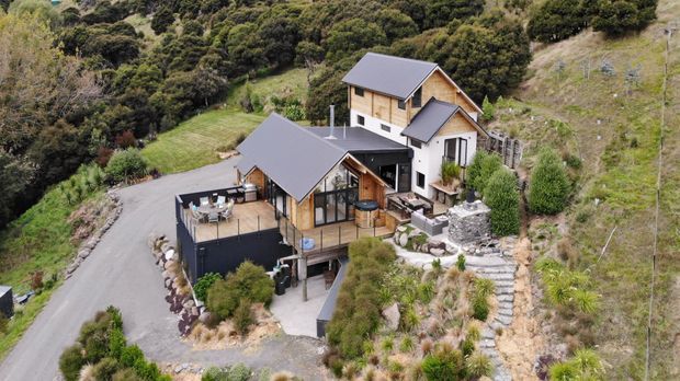Aerial view of a contemporary multi-level hillside home surrounded by dense native vegetation. The property features dark pitched roofs, timber and light-coloured exterior walls, extensive glazing, elevated decks and terraces, landscaped garden areas, and a paved driveway on a steep sloping site.