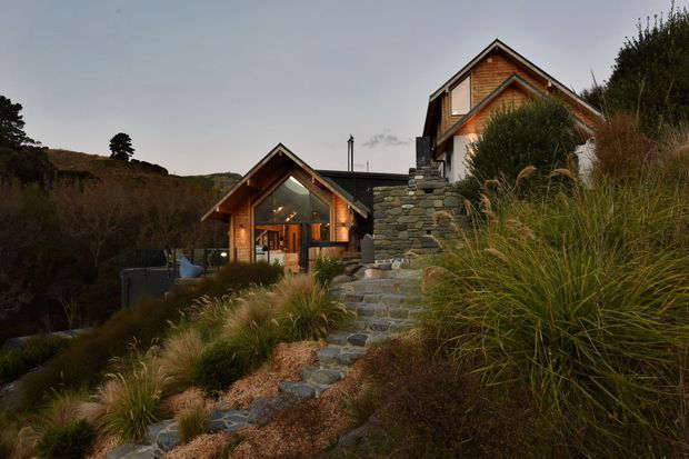 Exterior view of a pair of steeply gabled homes on a sloping, naturalistic site. The buildings combine dark timber cladding and exposed timber gable detailing with extensive stone retaining walls and steps. Warm interior lighting glows through the glazing, while native grasses and dense planting frame the approach.