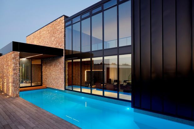 Contemporary multi-level residence with a strong geometric composition, combining warm exposed brick walls, dark metal cladding and extensive floor-to-ceiling glazing. A vivid illuminated swimming pool runs alongside a timber deck in the foreground, while warm interior lighting is visible through the glass.