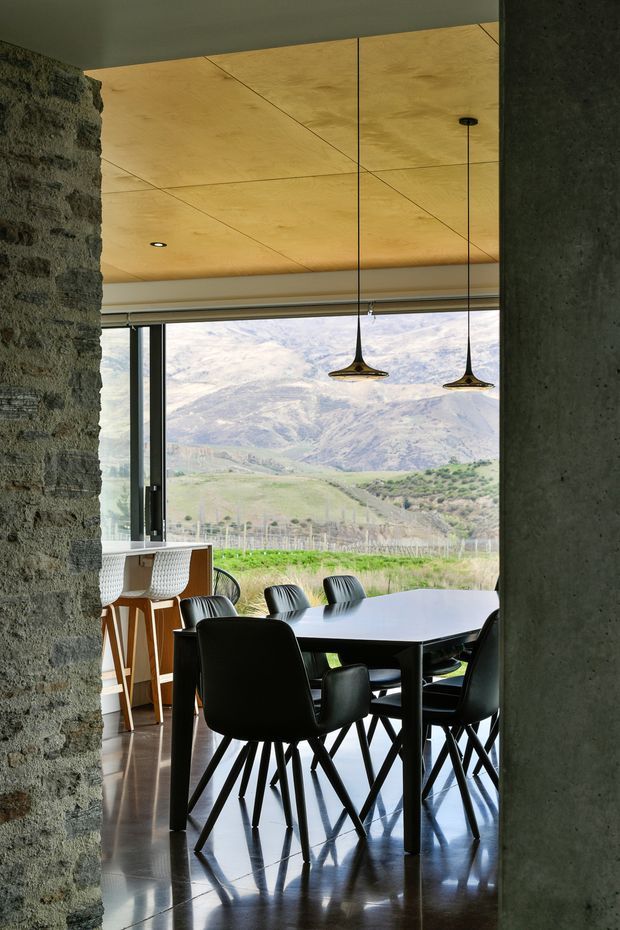 A refined dining area viewed through a rough stone and concrete opening. The room features a large dark dining table with black upholstered chairs, a timber-clad ceiling, polished dark flooring, and two sculptural pendant lights. Full-height glazing frames a rural mountain and vineyard landscape, while a compact timber-and-white kitchen or bar area is visible to the left.