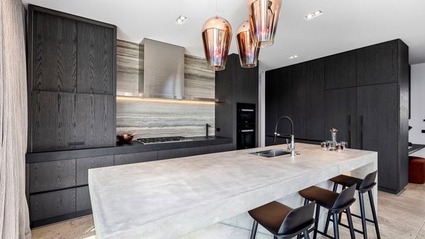 A contemporary kitchen featuring a large pale stone island with integrated seating, dark textured full-height cabinetry, a concealed rangehood and cooktop wall, stainless-steel appliances, and sculptural copper pendant lights. The space is bright, refined, and high-contrast, with timber-toned flooring and warm metallic accents.