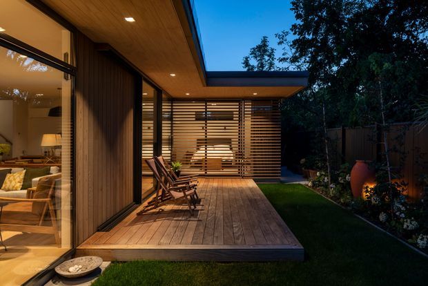 Twilight view of a contemporary timber-clad home opening onto a raised timber deck and sheltered courtyard. Floor-to-ceiling glazing reveals a warmly lit living room, while a horizontal timber screen encloses the covered outdoor area. The design features a strong indoor–outdoor connection, integrated roof overhangs, timber soffits, and landscaped garden edges.
