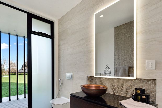 Contemporary bathroom with a floating dark vanity, copper vessel basin, mosaic tile splashback, large illuminated mirror, and light textured wall finish. A black-framed frosted-glass door opens toward a bright lawn outside.