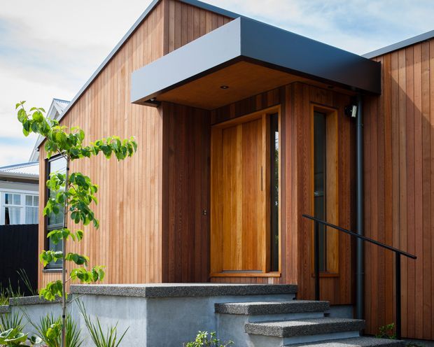 A modern residential entry facade with vertical timber cladding, a projecting dark flat-roof canopy, and a timber front door with narrow glazed sidelights. The entry is approached by several concrete steps with dark textured stone-like treads and a simple black handrail. A small landscaped garden and neighbouring building are visible around the entrance.