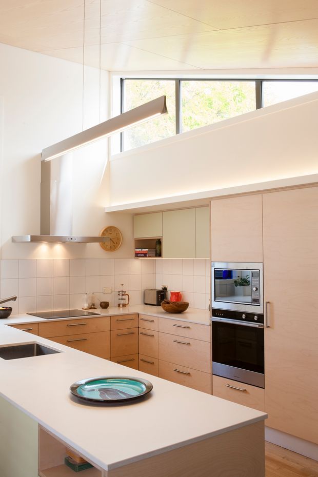 Bright contemporary kitchen with pale timber cabinetry, white tiled splashback, and a large white island. Integrated stainless-steel oven and microwave sit within full-height cabinetry, while a linear rangehood, open shelving, and broad clerestory windows provide a clean, airy fit-out. Light timber ceiling and warm timber flooring add natural warmth.