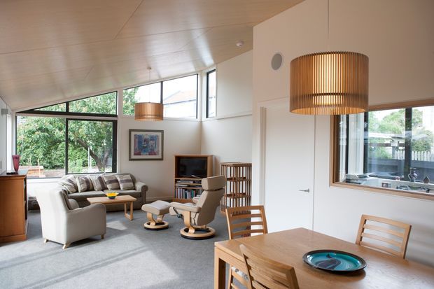 Bright open-plan living and dining area with a sloping timber-lined ceiling, extensive windows, pale walls, and grey carpet. The living zone contains sofas, lounge chairs, a television and shelving, while a timber dining table with matching chairs occupies the foreground. Large woven pendant lights and timber furniture add warmth to the contemporary interior.