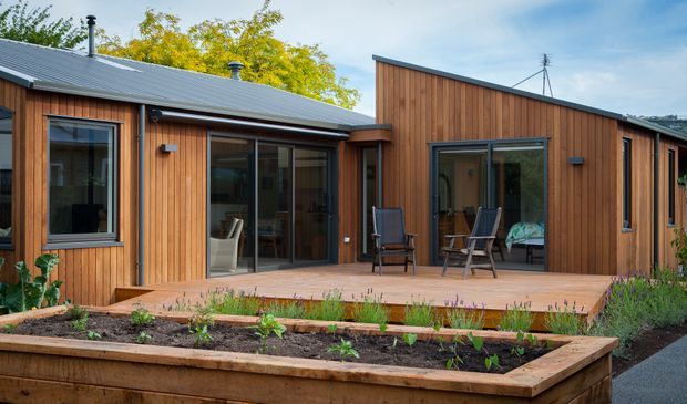 Single-storey timber-clad residential dwelling with a low-pitched metal roof and a taller asymmetrical gabled volume. Large glazed sliding doors open onto a broad timber deck, with raised garden beds in the foreground. The exterior has a warm, simple contemporary character and strong indoor–outdoor connection.