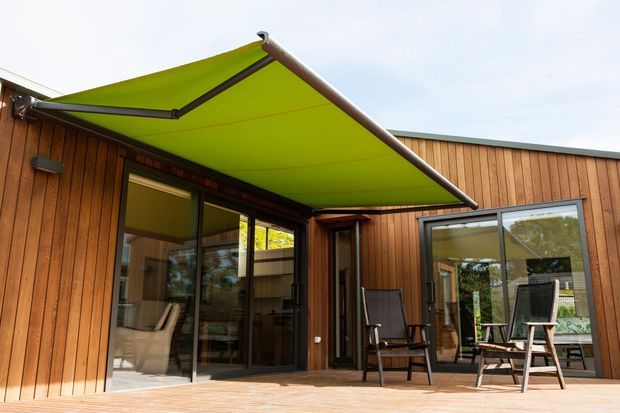 Single-storey timber-clad home opening onto a timber deck and sheltered outdoor sitting area. Large glazed sliding doors connect the interior to the deck, while a bright lime-green retractable awning provides shade over the patio. The view emphasizes the rear facade and indoor-outdoor connection.