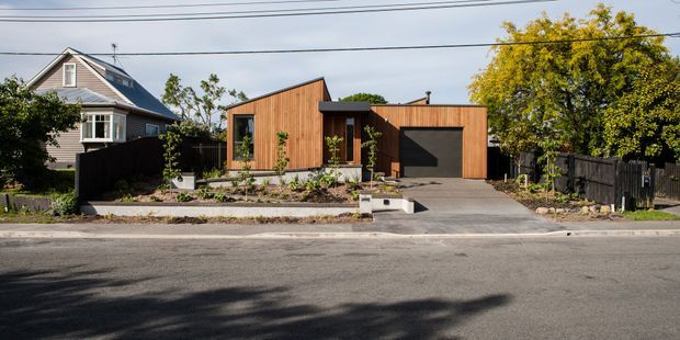 Street-facing view of a low, single-storey contemporary house with a compact angular form, warm vertical timber-clad walls, a dark garage door, and a simple concrete driveway. The landscaped front garden includes raised planting beds and a narrow entry path, while a larger traditional neighbouring house is visible to the side.