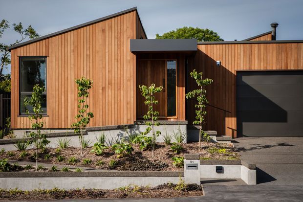 Front exterior view of a contemporary single-storey home with warm vertical timber cladding, angular pitched roof forms, dark-framed glazing, a recessed entry, and an integrated dark garage door. The house sits behind a landscaped garden and concrete retaining walls.
