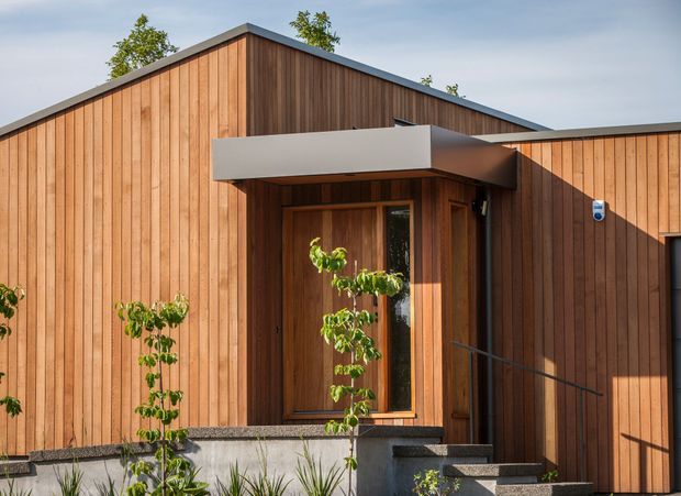 Exterior view of a contemporary single-storey building clad in vertical warm timber boards. A recessed timber entry with a projecting flat-roof canopy, dark metal roof edging, concrete steps, and simple landscaped planting define the facade.