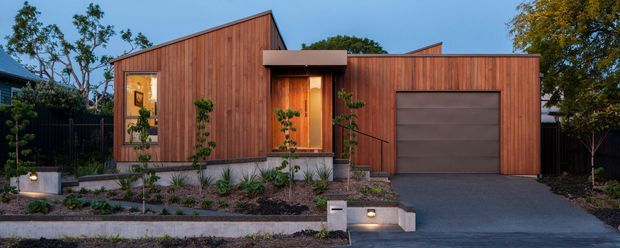 Front exterior view of a compact contemporary house with warm vertical timber cladding, angular pitched roof forms, a recessed timber entry door, and an integrated single garage with a broad grey sectional door. The landscaped frontage includes raised concrete garden beds, low planting, and a dark driveway, photographed at dusk.