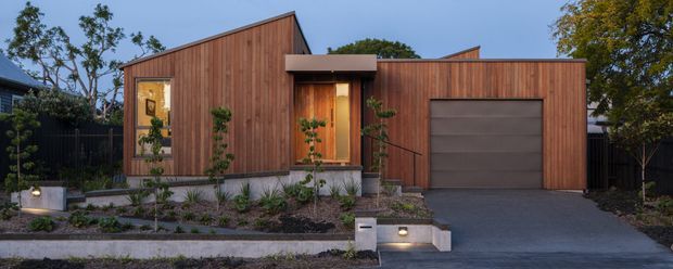 Street-facing exterior view of a compact contemporary home with a strongly articulated timber-clad facade. The building combines a steep asymmetric pitched roof over the main volume with a lower flat-roofed garage wing, warm vertical wood cladding, dark window and door frames, a recessed illuminated entry, and a wide grey garage door. A landscaped front garden with low planting, concrete retaining edges, and a driveway completes the setting.