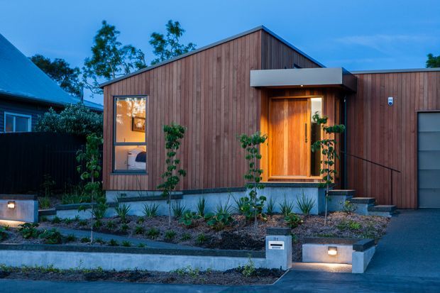 Exterior dusk view of a compact contemporary single-storey home. The facade is clad in vertical warm-toned timber boards, with a simple gabled roof, recessed timber entry door, projecting flat canopy, large window, concrete plinths and landscaped stepped garden beds leading to the entrance.