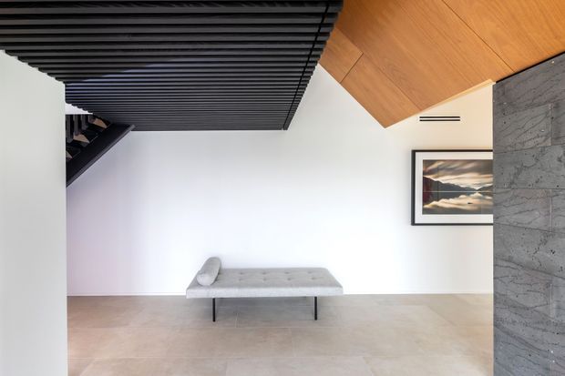 Minimalist interior with a low grey upholstered bench against a white wall. The room features large pale stone or concrete-look flooring, a dramatic black slatted ceiling and stair element, warm timber ceiling lining, and a textured grey stone feature wall with framed artwork.