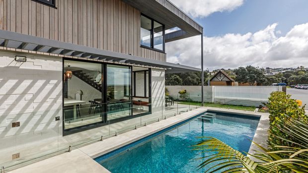 Modern multi-level residence with vertical timber cladding, exposed concrete block walls, extensive black-framed glazing, and a covered terrace opening onto a rectangular swimming pool. The pool terrace and contemporary architectural form dominate the view, with suburban surroundings visible beyond.