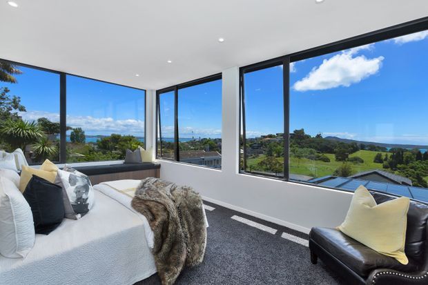 A bright contemporary living room with expansive corner glazing overlooking a green suburban landscape and distant ocean. The room features a white slipcovered sofa, black leather armchair, textured throw, dark carpet, recessed ceiling lights, and crisp white walls with slim dark window frames.