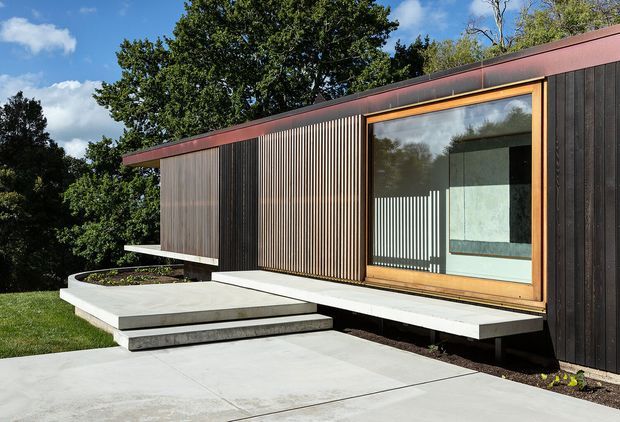 Exterior view of a compact, single-storey modern building set within a leafy landscape. The facade combines dark vertical timber cladding with finely spaced timber screening, a broad timber-framed glazed opening, and a low reddish-brown metal roof edge. A raised concrete terrace and stepped concrete entry path lead to the building, with the interior visible through the glazing.
