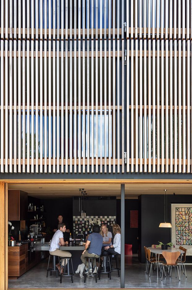 A contemporary two-storey facade with a dramatic vertical timber batten screen over large glazed openings. The lower level is an open-plan interior featuring a dark kitchen and bar area, central island seating, exposed dark structural columns, timber-lined ceiling, and an adjacent dining space. The upper facade combines warm timber framing with closely spaced vertical battens, allowing filtered views through the glazing.