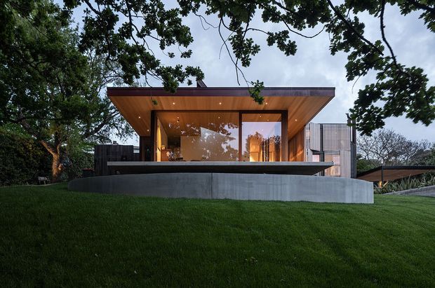 Exterior view of a low-slung contemporary house set within a landscaped, tree-lined site. The composition highlights a projecting flat roof with warm timber soffit, extensive floor-to-ceiling glazing, dark upper cladding, and a raised concrete terrace or retaining wall overlooking a sloping lawn.