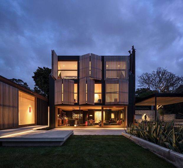 Twilight exterior view of a contemporary multi-storey residence arranged around a landscaped courtyard. The dark facade features extensive vertical timber or metal screening, large glazed openings, warm interior lighting, and projecting louvred panels. A lower side pavilion and covered outdoor structure frame the central house, with concrete paving and raised garden beds in the foreground.