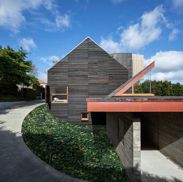 Exterior view of a contemporary gable-roofed building clad in horizontal weathered timber boards. A curved paved walkway leads beside landscaped groundcover to a recessed garage or covered entry, with a prominent rust-coloured metal roof or canopy element projecting across the foreground. The composition features strong geometric forms, a pitched gable facade, and contrasting vertical metal screening under a bright blue sky.