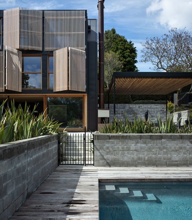 Contemporary multi-level residential exterior with dark cladding, extensive vertical timber screening, and warm timber-framed glazing. A timber pool deck and swimming pool occupy the foreground, while raised garden beds with architectural planting, concrete block retaining walls, a metal gate, and a covered outdoor structure frame the property. Bright daylight and strong linear shadows emphasize the facade and landscape geometry.