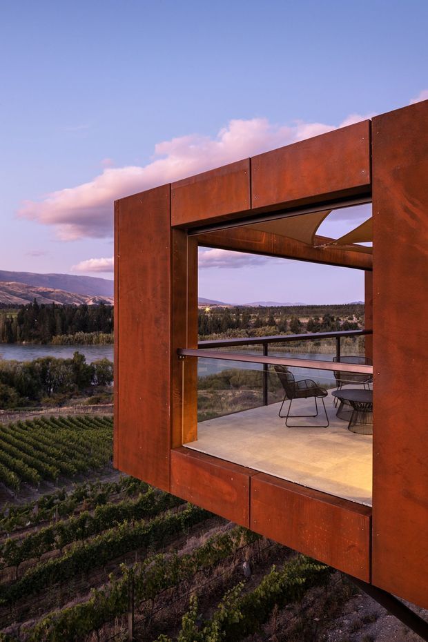 A dramatic elevated architectural terrace or viewing platform framed by large rust-coloured weathering steel panels and exposed structural beams. The open-sided space contains a small outdoor dining setting, with expansive glazing and views across a vineyard, lake or river, forest, and surrounding hills at sunset. The image strongly emphasizes the cantilevered steel construction and scenic rural landscape.
