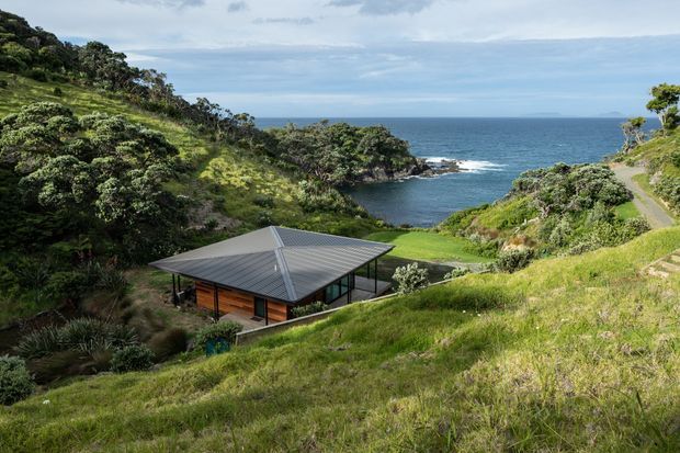 A low, modern house with a dark hipped metal roof and timber-clad walls is nestled into a steep, lush coastal hillside. The property overlooks a rocky cove and open ocean, with a narrow driveway or path descending along the right side. Dense native vegetation and grassy slopes surround the building.