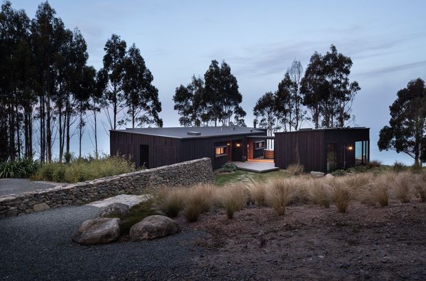 A low, single-storey contemporary house composed of dark timber-clad pavilion-like volumes, set within a rugged landscaped site. A recessed, warmly lit entry or connecting space sits between the volumes, with a small deck, stone retaining wall, gravel ground, boulders, ornamental grasses, and tall eucalyptus trees surrounding the building. The image is photographed from outside at dusk, emphasizing the dark facade against the pale sky and warm interior lighting.