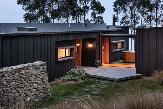 Exterior view of a low-slung contemporary cabin-like home clad in dark vertical timber boards. The building has a shallow pitched metal roof, warm-lit windows and an open timber-framed doorway leading to a sheltered deck with a round outdoor spa or bath. A rough stacked-stone wall, stepping-stone path and native grasses integrate the dwelling into a natural, wooded setting.