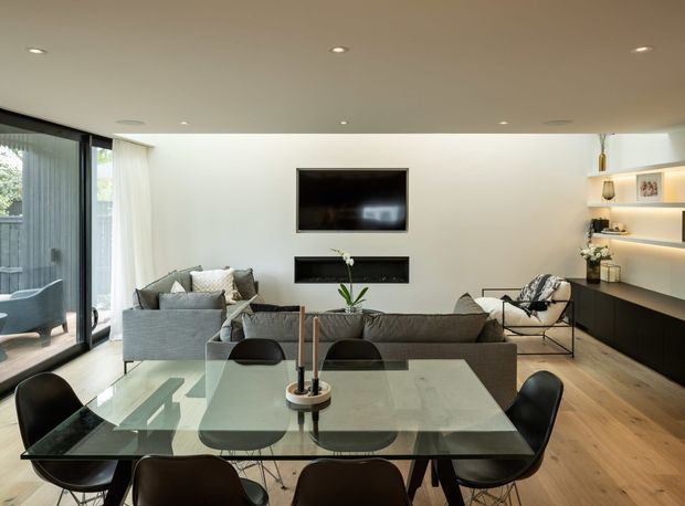 Open-plan contemporary living and dining area with a large glass dining table and black chairs in the foreground, a grey sectional sofa facing a wall-mounted television and linear fireplace, and built-in dark cabinetry with illuminated display shelving. Floor-to-ceiling glazing with sheer curtains opens to an outdoor area, while light timber flooring and a recessed ceiling create a bright, refined interior.