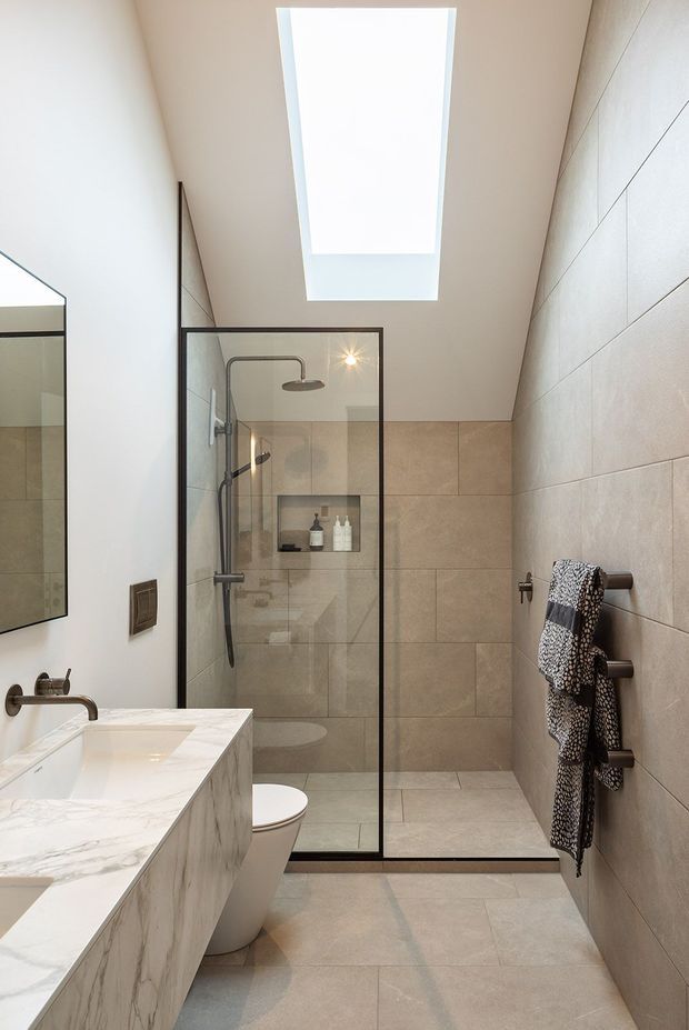 A bright contemporary bathroom with a walk-in glass shower enclosure, large-format beige wall and floor tiles, a white marble-look vanity with integrated basin, wall-mounted tapware, and a skylight providing abundant natural light. Dark metal shower framing and fixtures add contrast.