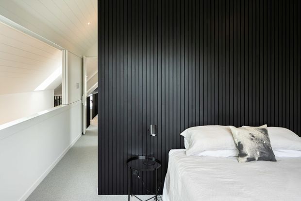 A bright contemporary bedroom viewed from beside a hallway or landing. The room features a white bed with layered white bedding and a grey accent cushion against a dramatic black vertical timber-panel feature wall. A small black bedside table and wall-mounted reading light sit beside the bed. To the left, a white-lined corridor or mezzanine-like passage is visible with a sloping ceiling, pale carpet, and skylight-like natural light.
