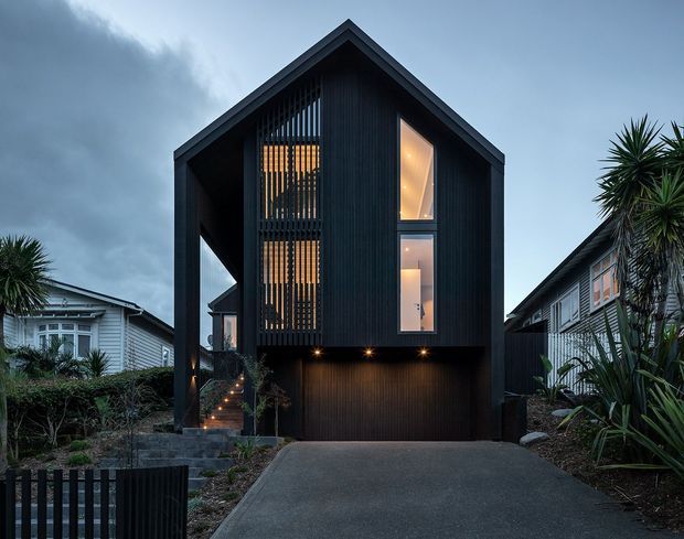 Front exterior view of a narrow, contemporary multi-storey house with a steep gabled roof, dark vertical cladding, dramatic recessed entry stairs, tall glazed openings, and an integrated garage at driveway level. The home is set among established neighbouring houses and subtropical planting, photographed in subdued evening light.