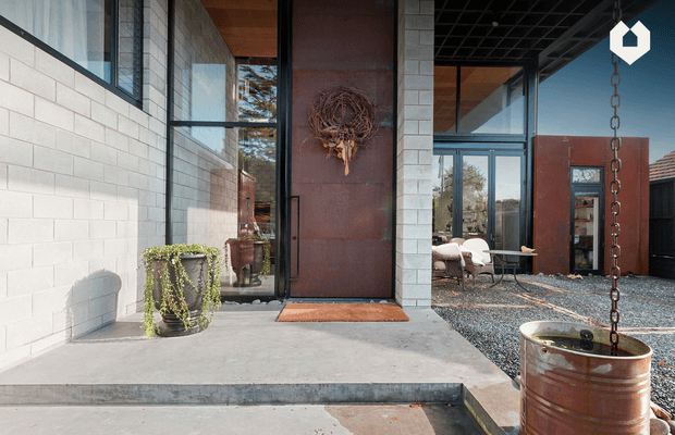 Contemporary industrial-style residential entry viewed from the exterior, featuring pale concrete masonry walls, expansive black-framed glazing, a tall rusted steel pivot door, exposed timber soffit, and a gravel courtyard. A suspended rusted metal planter hangs beside the entry, while a seating area is visible through the glazing.