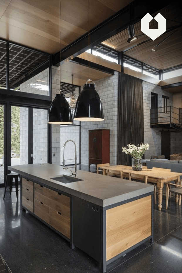 A dramatic open-plan kitchen and dining space with a large timber-and-black island, dark stone benchtop, undermount sink and gooseneck tap. Black pendant lights hang above the island. The room features polished dark flooring, pale concrete block walls, timber ceilings, exposed black structural framing, large windows, and a raised mezzanine or landing with black metal balustrades. A timber dining table and chairs sit behind the island.