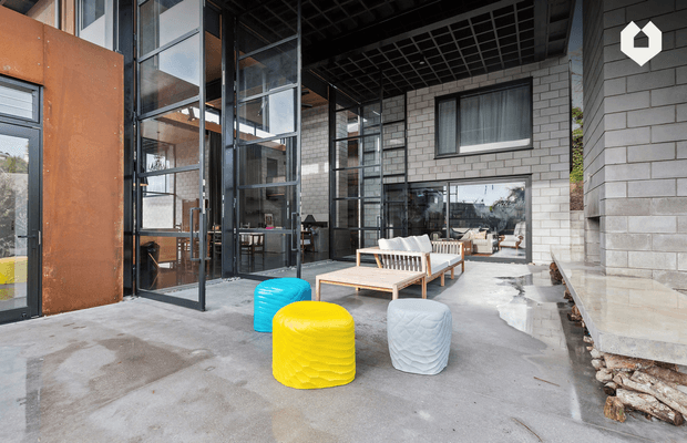 A contemporary covered outdoor living area beside a modern building, with a raw concrete floor, exposed grid ceiling, large black-framed glazed openings, grey concrete block walls, and a rust-toned metal-clad section. A timber-framed lounge setting with white cushions is accompanied by bright yellow, turquoise, and white sculptural stools. The space connects visually to the building interior through extensive glazing.